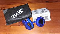 Blue Snark SN-1x clip-on tuner, black carrying pouch with white Snark logo, and instruction manual placed on wooden surface.