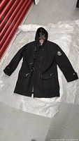 Full view of the black Tommy Hilfiger men's wool coat laid flat, showing toggle button closure, attached hood, and front pockets with blue trim.