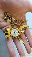 Hand holding gold-tone Golden Snitch pendant open showing small pocket watch with Roman numerals.