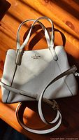 White leather Kate Spade purse with dual handles and detachable strap lying on wooden surface showing full front view of the bag.