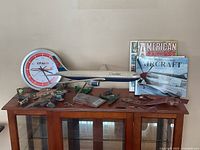 Image shows a CP Air branded wall clock, Canadian Airlines model airliner, several green and red biplane models, broken wooden airplane parts, and three aviation books arranged on a wooden table.