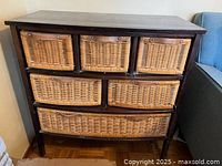Front view of wooden cabinet with seven natural wicker baskets as drawers, showing dust and surface wear.