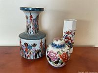 Three ceramic vases on a wood surface: large with crack repaired, small ginger jar style, and white hexagonal vase with floral design