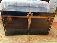 Black metal chest with brass hardware viewed from front on carpet.