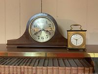 Wooden mantel clock and brass carriage style clock on shelf above books.