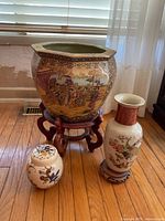 Photo showing all three ceramic decorative pieces on hardwood floor near curtain and window: large planter on wooden stand, tall vase on wooden base, and small covered jar.