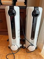 Photo showing two identical DeLonghi oil-filled radiator heaters placed side by side, cables visible, slight dust and scratches on surfaces.