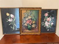 Three framed floral paintings placed side by side on a table showing a variety of flowers in different frame styles and colors.