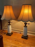 Pair of working vintage table lamps with white ceramic bases, brass accents, square wood and brass bases, and beige scalloped fabric shades. Lamps are lit to show warm glow and adjustable light intensity.