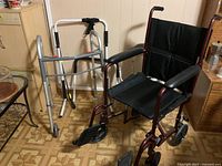 Photo showing a burgundy manual wheelchair, a white walker, and a silver walker placed on a patterned floor near wood-paneled walls.