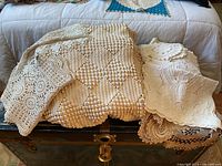 Stack of crocheted and needlepoint linens including a diamond pattern crocheted blanket and lace doilies on a wooden chest