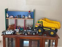 Wide view of used toys including large yellow Tonka dump truck with rust, multi-color wooden shelf, small toy cars, and collectibles arranged on dark wood cabinet.