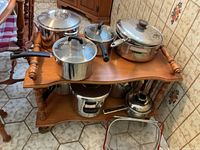 Multiple stainless steel pots and saucepans with lids displayed on a wooden trolley, showing various sizes and handle types