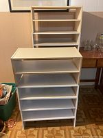 Photo of two white wooden shelving units with multiple shelves shown side by side, one slightly taller than the other, placed on patterned floor.
