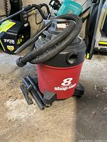 Shop-Vac 8 gallon wet/dry vacuum cleaner with hose and several attached cleaning tools, shown from slightly different angles on concrete floor.