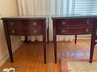 Pair of reddish-brown wooden side tables with two drawers each. Brass ring pull handles and tapered legs visible. Positioned on hardwood floor with natural light from window.