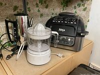 Photo showing black Ninja Foodi multi-cooker, white electric citrus juicer and stainless steel coffee percolator on countertop