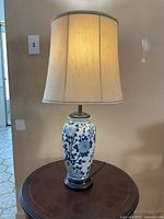 Full view of the lit table lamp on a wooden surface showing its porcelain vase base with blue floral design and light fabric shade.
