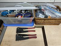 Photo of three manual staple guns with red handles and a metal bevel square laid on a wooden surface. Also visible are plastic containers and woven baskets holding numerous small tools and parts.