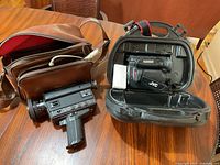 Photo of vintage rectangular black film camera with pistol grip and brown leather carrying case on wooden table.