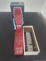 Red Merlin handheld electronic game standing open next to original box showing inside of device and speaker panel.