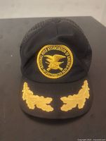 Front view showing circular embroidered NRA logo with gold eagle and crossed rifles, and gold oak leaf embroidery on brim.