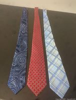 Three ties laid out on a surface, showing front and patterns: dark blue paisley, red geometric, light blue plaid.