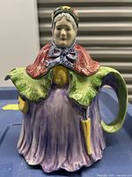 Front view of the ceramic tea pot showing the woman in colorful dress with bonnet, blue scarf, green collar, purple skirt, and a yellow umbrella handle.