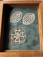 Framed embroidery showing three vintage lace pieces on dark textured fabric inside a wooden frame.
