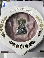 Front view of the circular white and gold-decorated wall hanging with a metal religious sculpture inside over pink fabric.