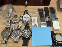 Overview of watches and watch bands arranged on a surface, including silver, black, and gold tones, with some watches detached from bands and packaged Timex bands.
