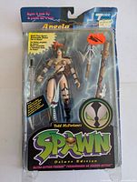 Front view of Todd McFarlane Angela figure in original blister packaging showing figure, accessories, and card backing with visible wear and cracks