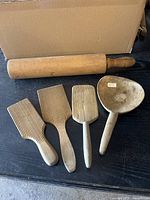 Photo showing one wooden rolling pin missing one handle, three wooden butter paddles with grooved surfaces, and one wooden butter spoon on a dark surface
