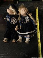 Photo of boy and girl porcelain head marionette dolls laying side by side showing their full bodies dress in blue velvet with plaid details and cream-colored shoes, with a measuring tape showing approximately 12 inches tall.