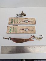 Four vintage fishing lures shown with two hex spinners on original cards, one unbranded spinner, and a Johnny Green brass trolling spoon with hook and beads, placed alongside a ruler for scale.