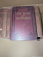 Front cover of one book in set showing title 'The Book of Knowledge' with gold embossing on maroon hardcover with ornamental corner details.