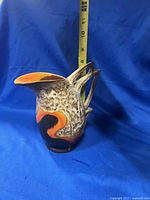 Side front view of the Art Deco ceramic pitcher showing the textured surface, abstract orange and black decoration, flared spout and double handles, photographed against blue backdrop with a ruler for scale (approximately 8 inches height).