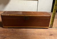 Closed view of the long rectangular wooden cigar box with a metal clasp and slight wear marks on the surface.