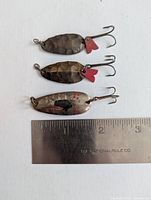 Top view of three fishing spoons with hammered and polished metal finishes, two with red tail attractors, shown next to a ruler for scale