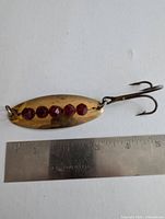 Close up photo of a brass fishing lure with six red circular reflective spots and attached treble hook next to a ruler showing size approx. 3.75 inches