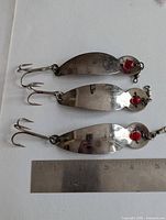 Photo showing three Gibbs One Eyed Wiggler size 4 metal fishing lures with treble hooks and red eye decoration next to a ruler for size reference.