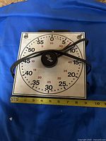 Top view of vintage GraLab timer showing dial markings, large black knob with logo, and attached power cable, placed on blue fabric background with measurement tape beside it showing approximately 10 inches in width.