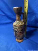 Vertical image of the Greek lekythos style pottery pitcher with measuring tape against a blue backdrop, showing it is approximately 7 inches tall. Visible black-figure design and aged ceramic surface.