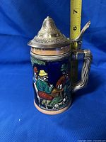 Front view of the vintage beer stein showing a colorful painted scene of people sitting at a table, with a pewter lid and a measuring tape showing approx 6.5 inches tall.