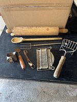 Vintage kitchen utensils arranged on black surface including wooden rolling pin, wooden spoon, meat fork, potato masher, tomato slicer, can opener, pie crust tool, tin scoop.