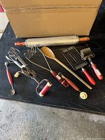 Photo shows 8 vintage kitchen utensils laid out on a black surface with cardboard box in background. Utensils have red handles and include rolling pin, hand mixer, potato masher, wooden spoon, melon scooper, tomato slicer, pastry roller, and pie crust tool, with visible wear and two broken wires on tomato slicer.