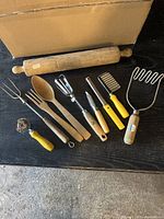 Photo of 9 vintage kitchen utensils including wooden rolling pin, pie crust tool, meat fork, salad tongs, hand mixer, cheese slicer, potato masher, potato peeler, and jar lid opener.
