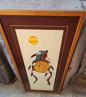 Framed S.Sabourin artwork titled 'Medicine Shield Clan' showing bear with decorated shield and orange sun against a neutral background framed with maroon mat and wooden frame.