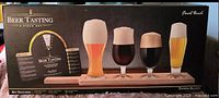 Front view of the box showing four beer glasses filled with different beers positioned on wooden paddle with 'Beer Tasting 6 Piece Set' title.