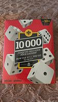 Front view of the sealed dice game "10,000" box showing the title and dice images on a red background.